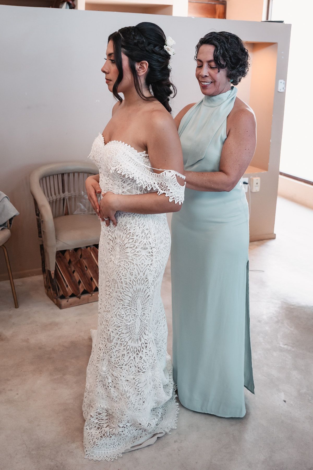 Bridesmaid in mint green dress helps bride fasten her white lace wedding gown before the ceremony.