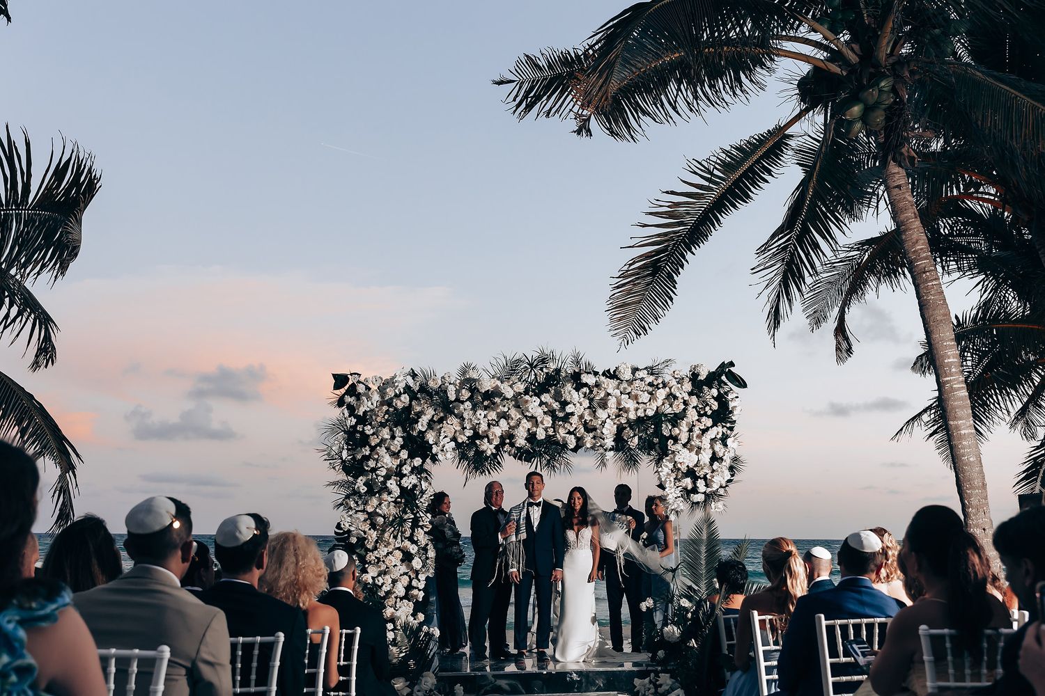 Wedding guests dance and celebrate during an outdoor sunset reception under palm trees.