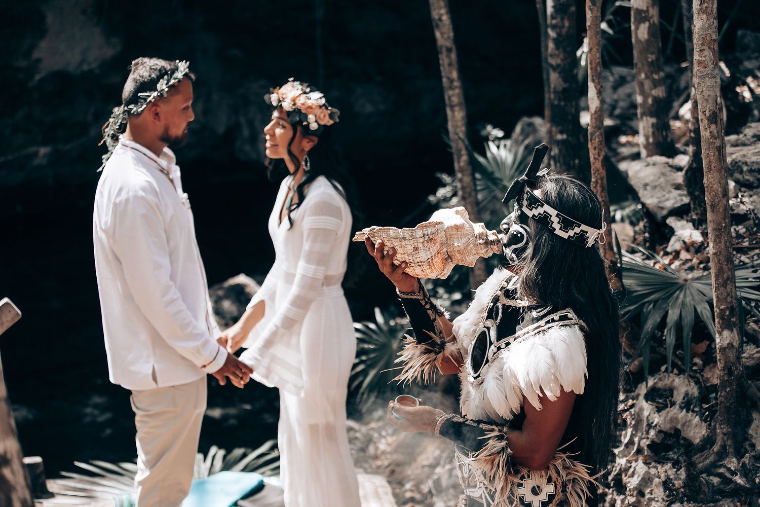 People in white attire gather for a traditional ceremony among trees and natural surroundings.