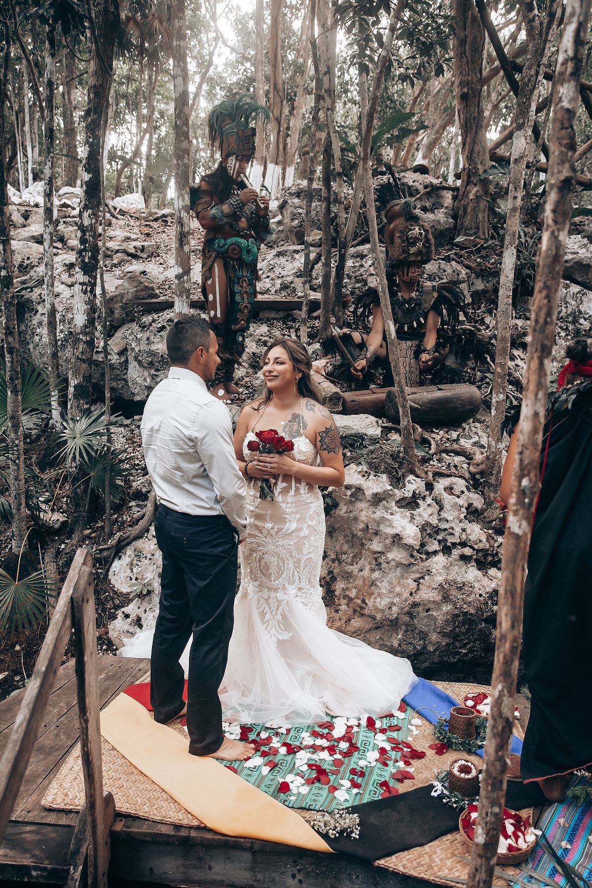 A couple celebrates their wedding ceremony in a rustic woodland setting with scattered rose petals and natural decor.