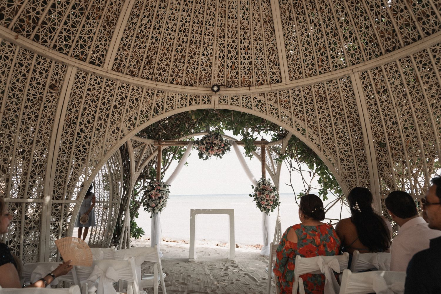 Elegant beach wedding ceremony under ornate white lattice gazebo with ocean view.