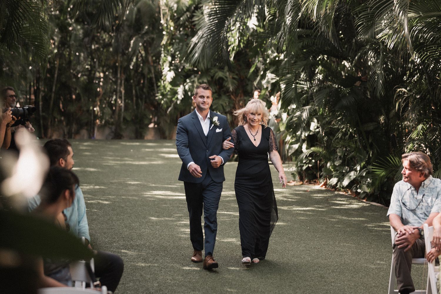 Groom escorts elegantly dressed woman down outdoor wedding aisle surrounded by lush tropical palm trees.