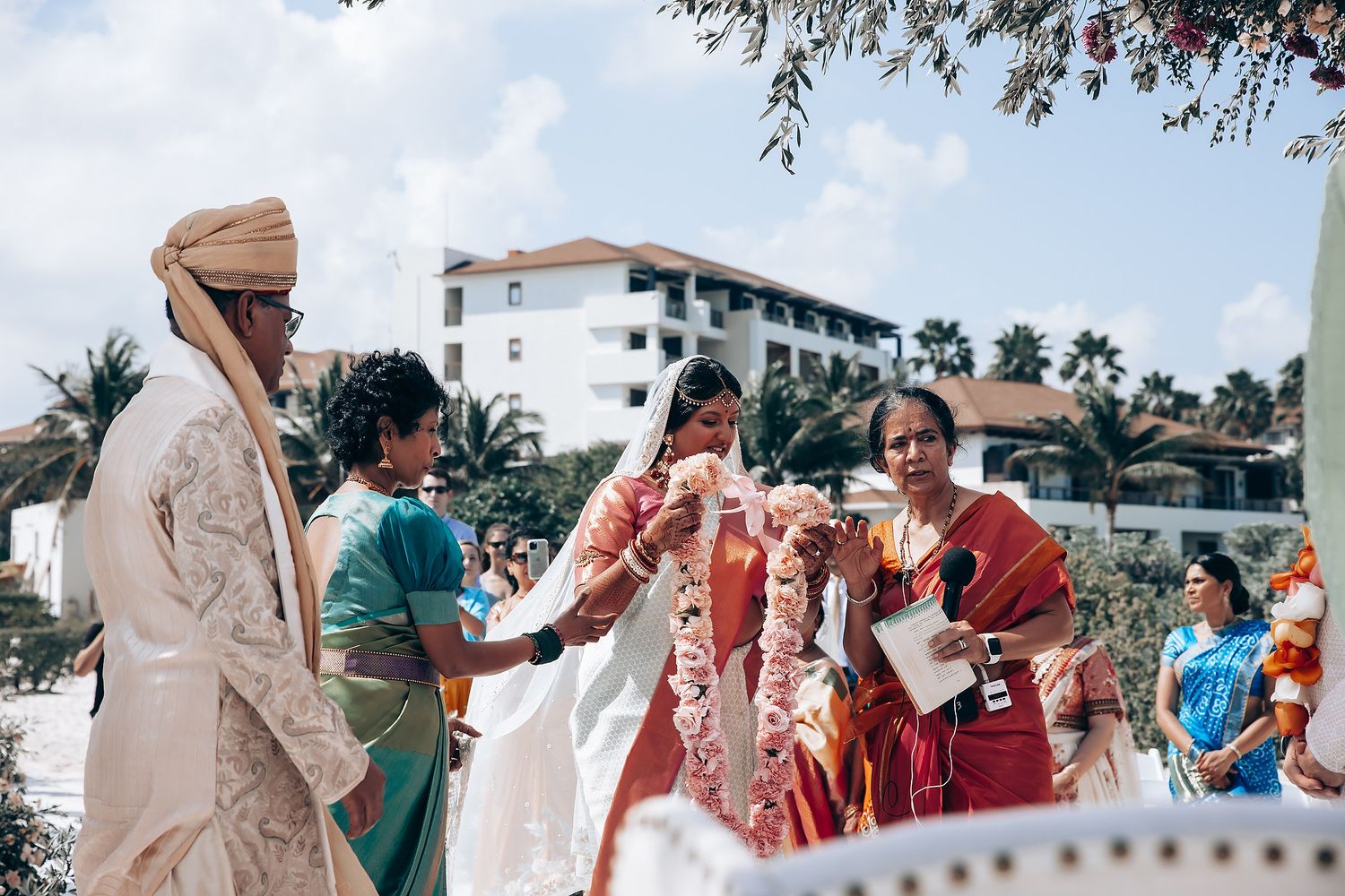 A traditional Indian wedding ceremony taking place outdoors with tropical architecture in background.