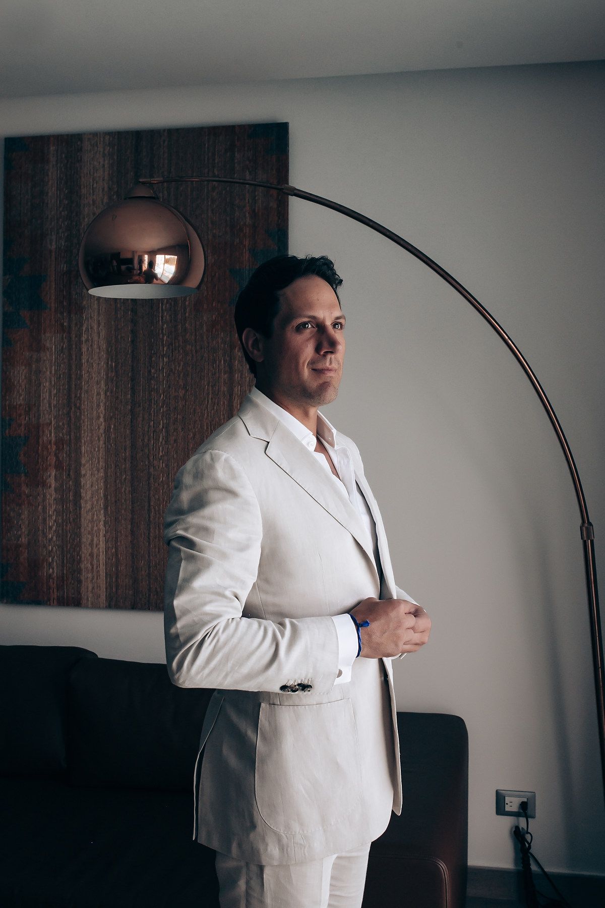 A person wearing a crisp white suit stands in a modern room with an arched floor lamp and dark wooden wall panel.