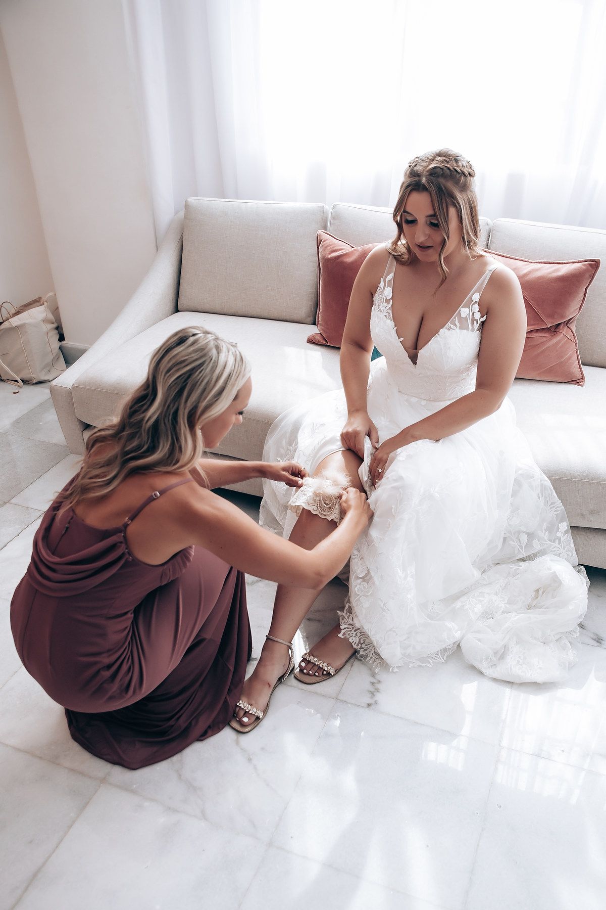 Bridesmaids in burgundy dresses help with wedding dress preparations in a bright white room.