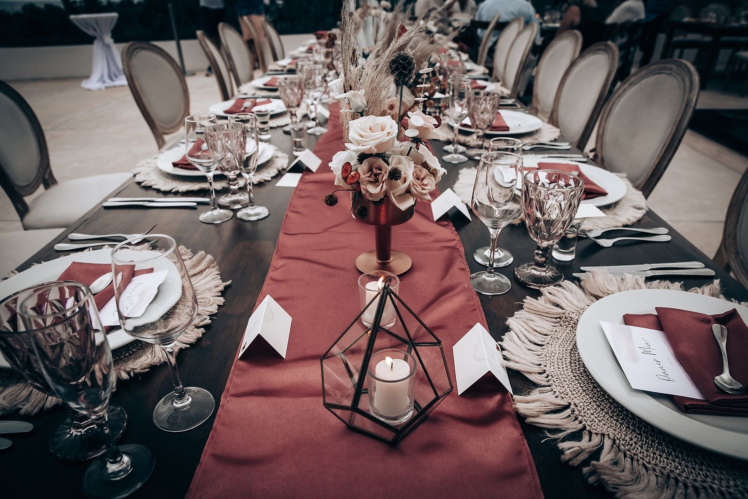 Elegant wedding reception table setting with burgundy accents and crystal glassware in dim lighting.