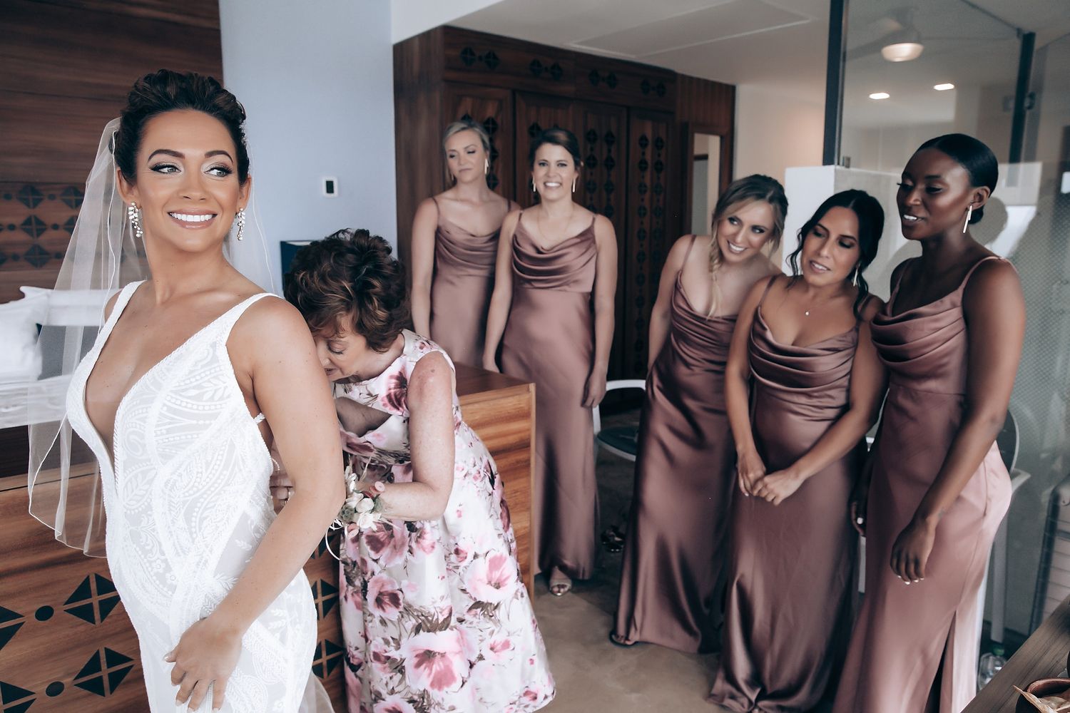 Bridesmaids in mocha brown dresses gather around the bride in white for pre-wedding photos at an indoor venue.