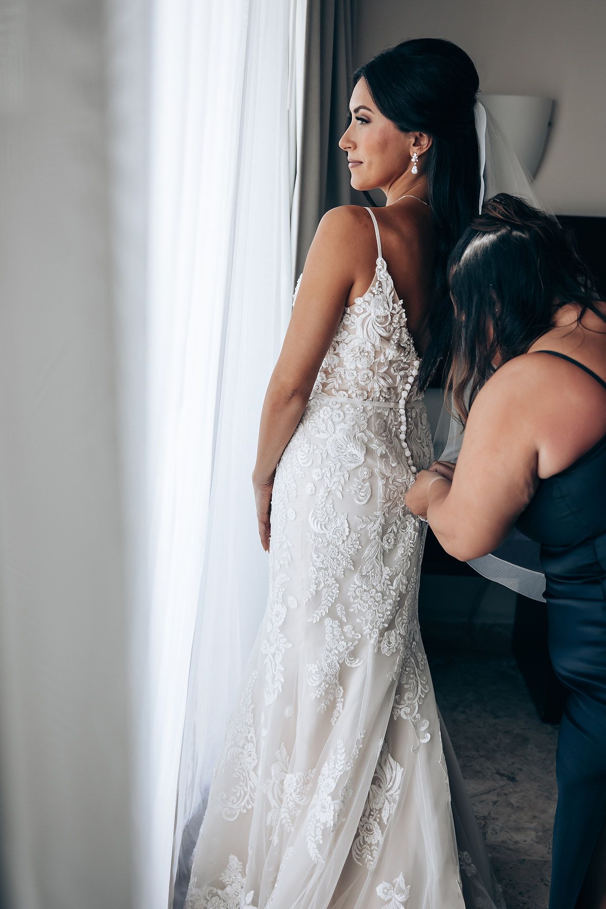 A bride in an elegant white lace wedding gown stands by a window in a getting ready room.