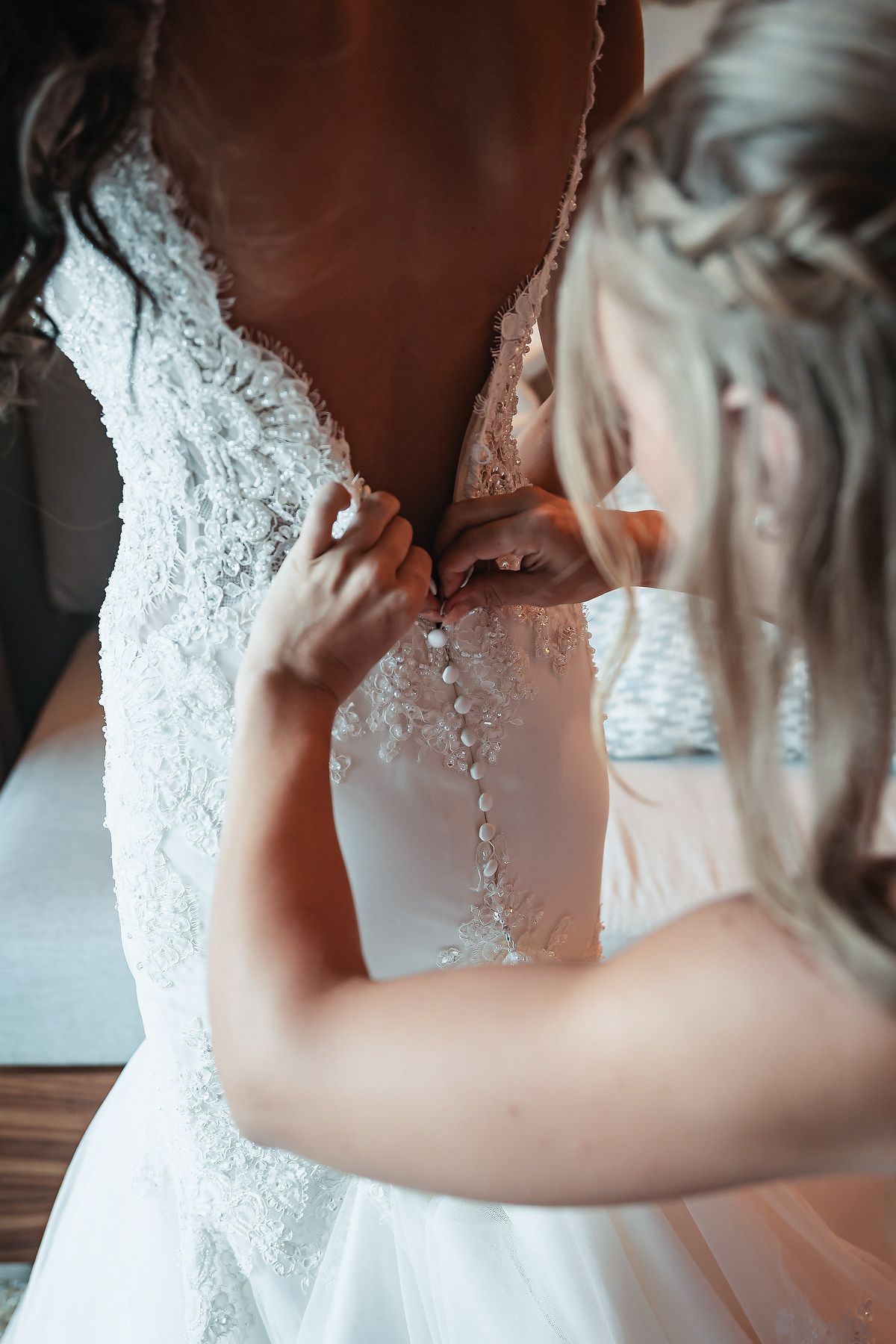 Bridesmaid fastening pearl buttons on back of elegant lace wedding dress during bridal preparation.