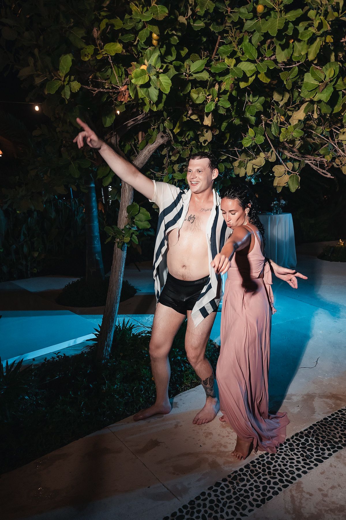A man and woman dancing outdoors at night near a pool, surrounded by tropical greenery and blue lighting.