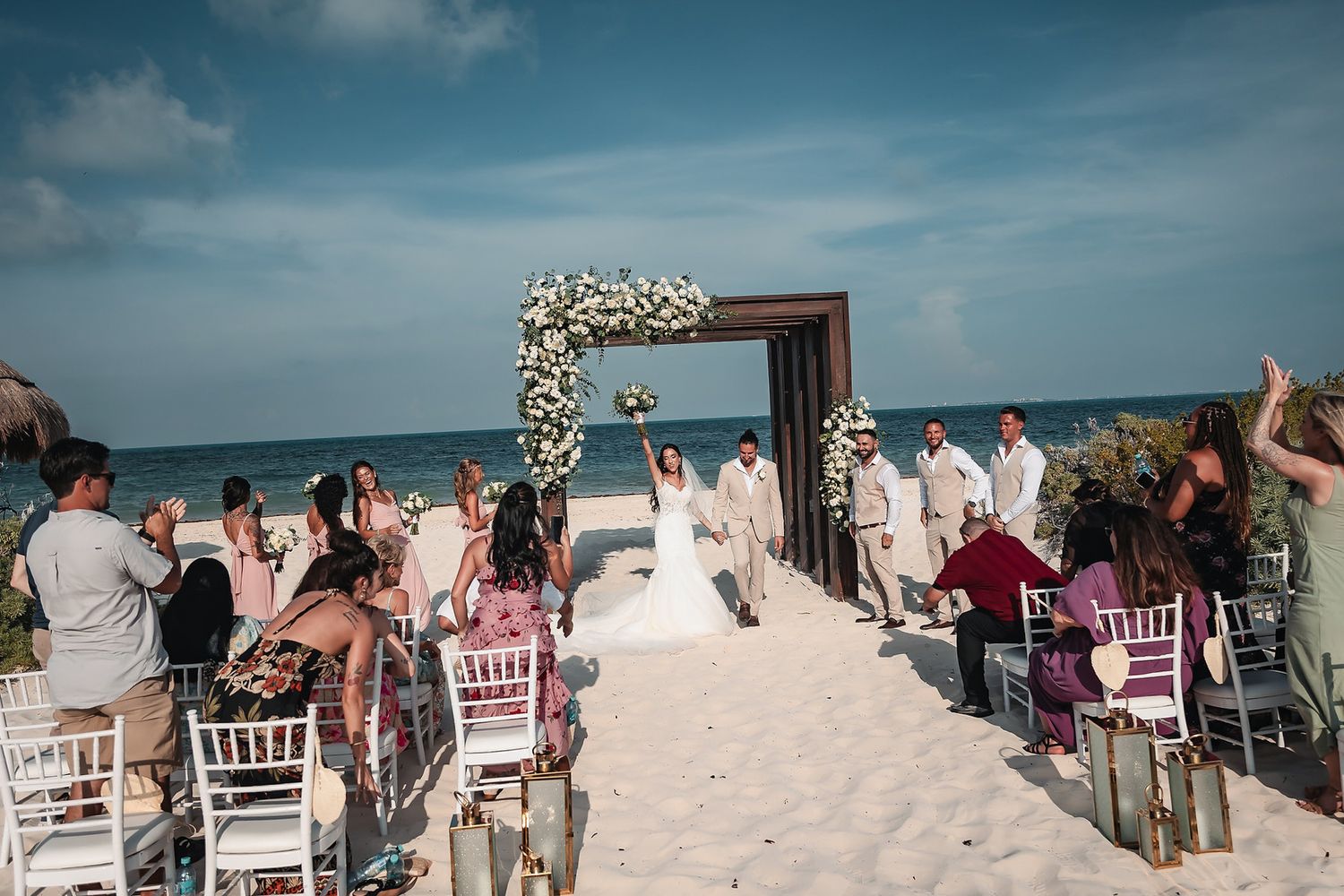 Romantic beach wedding ceremony with floral arch, white chairs, and guests on white sand with ocean backdrop.