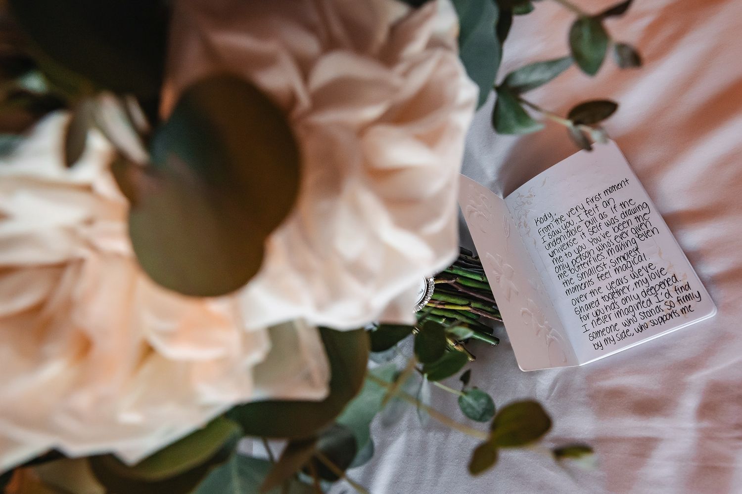 Bridal bouquet with blush peonies, eucalyptus, and a handwritten vow card on white fabric.