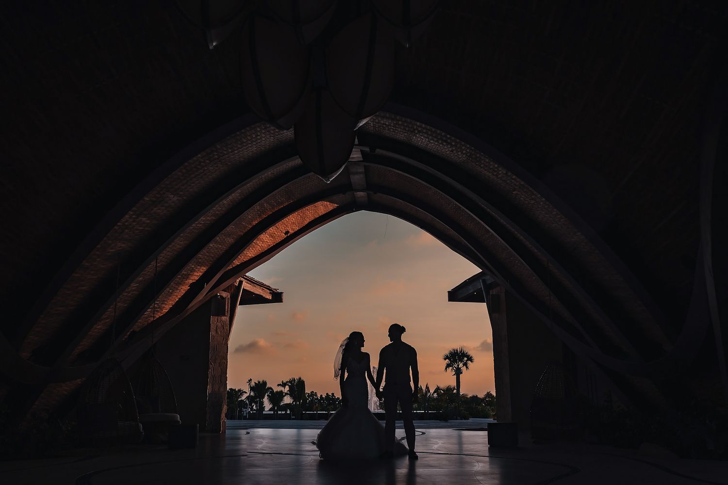 Silhouette of a couple holding hands framed by an arched structure at sunset with palm trees in the background.