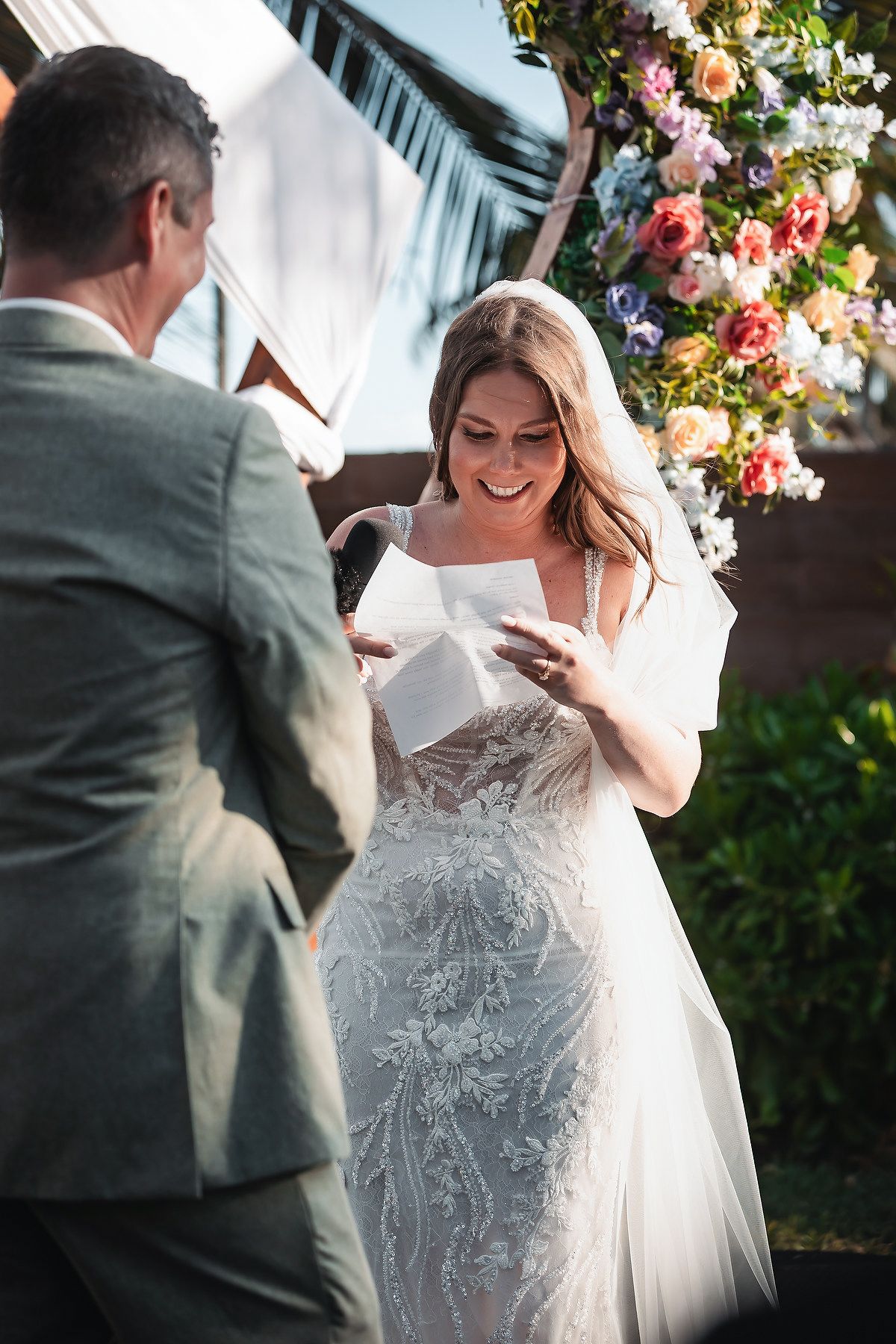 Smiling bride in lace gown reads vows from card at outdoor wedding ceremony with colorful floral arch.