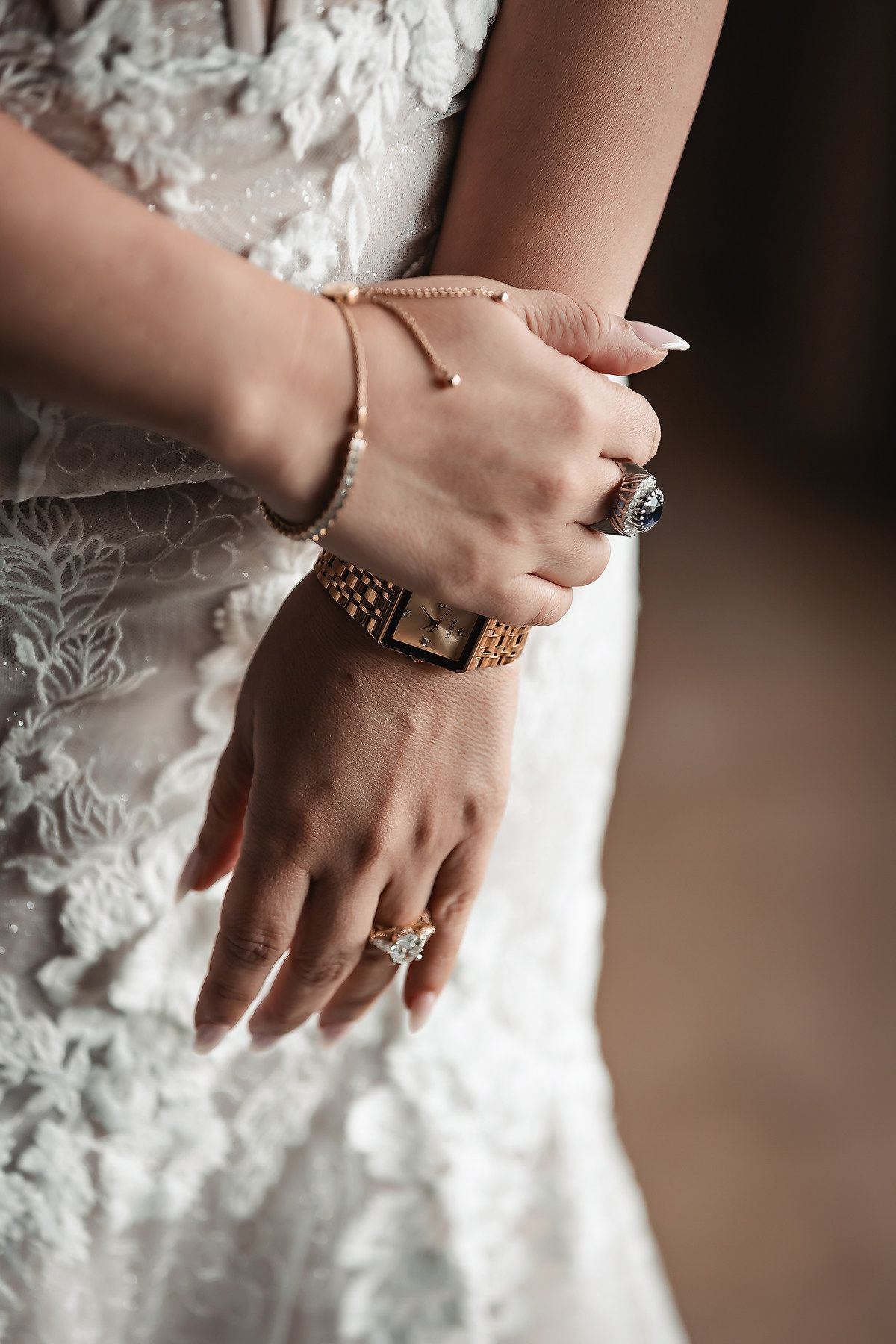 Bride wearing a diamond engagement ring, gold watch, and delicate bracelet against a white lace wedding gown.
