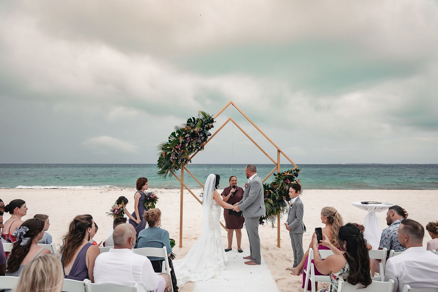 Couple exchanging vows at beach wedding under geometric copper arch with ocean backdrop and seated guests.