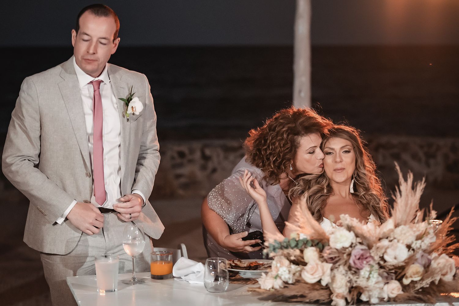 Bride and groom at outdoor beach wedding reception table with floral arrangements, as a guest shares a kiss on the cheek.