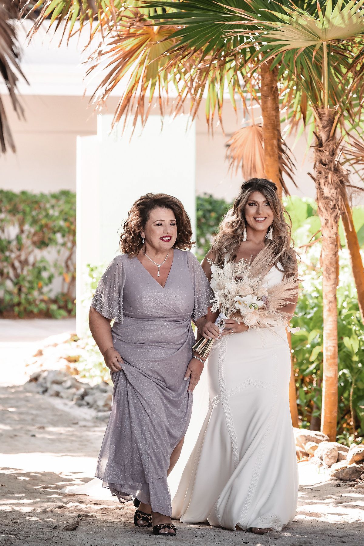 Bride in white gown walks with woman in lavender dress near palm trees at outdoor wedding venue.