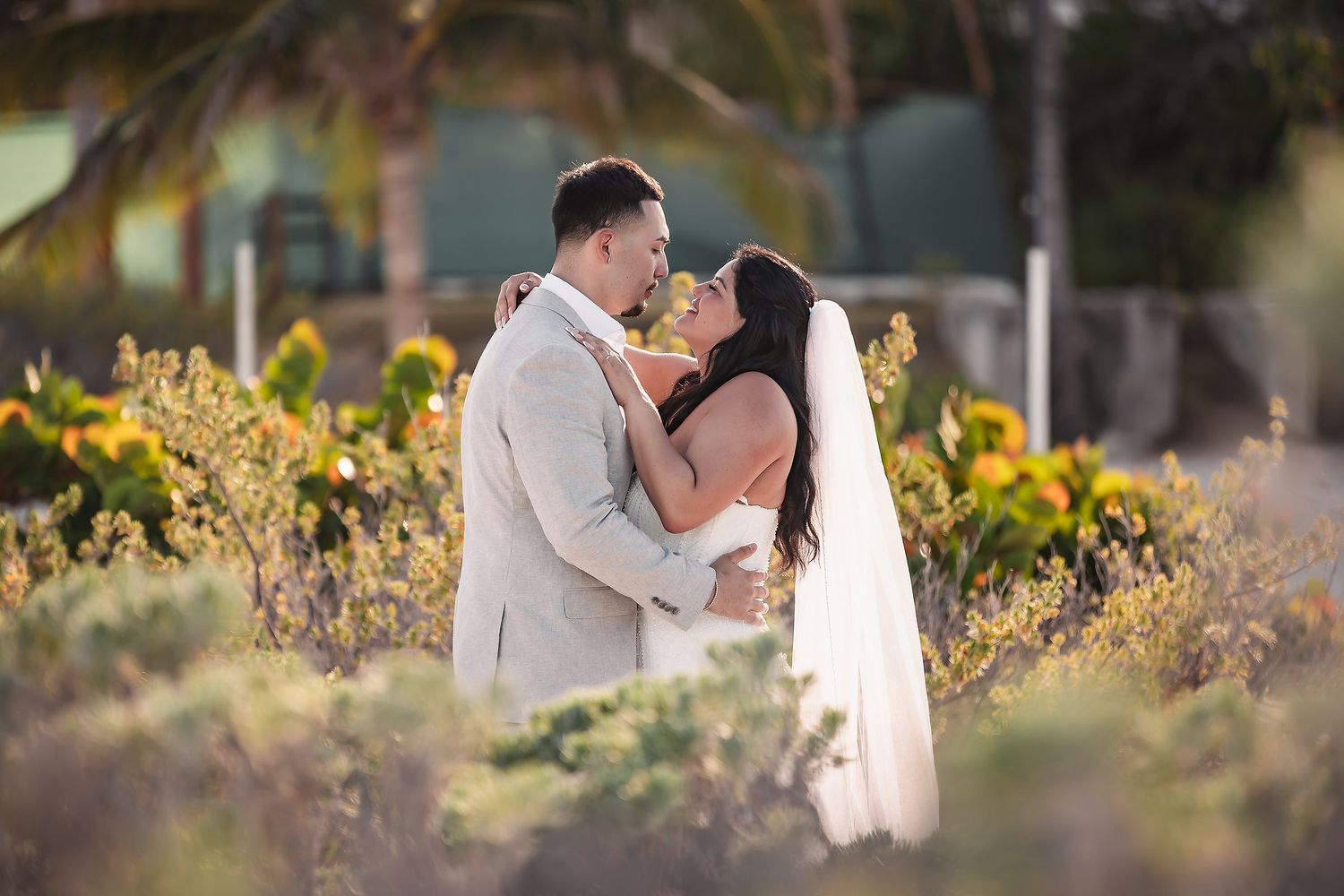 Bride and groom sharing a romantic almost-kiss embrace surrounded by lush tropical garden greenery at golden hour.