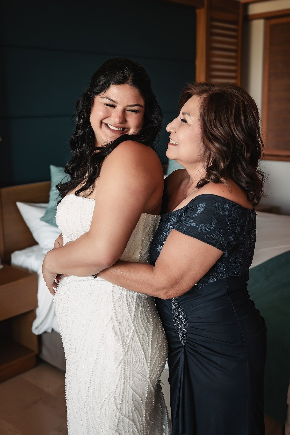 Bride in white lace gown shares joyful hug with mother in navy dress during wedding preparations.
