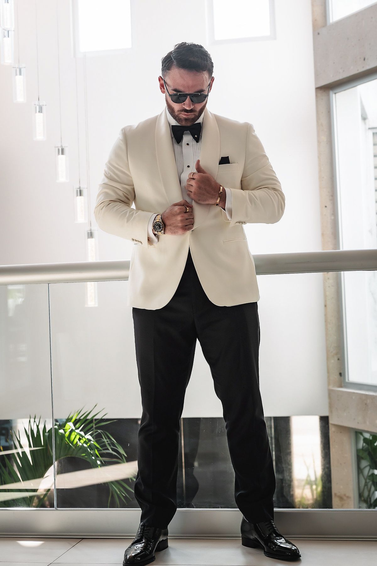 A well-dressed man in a white tuxedo jacket, black bow tie, and trousers adjusts his suit while wearing sunglasses indoors.