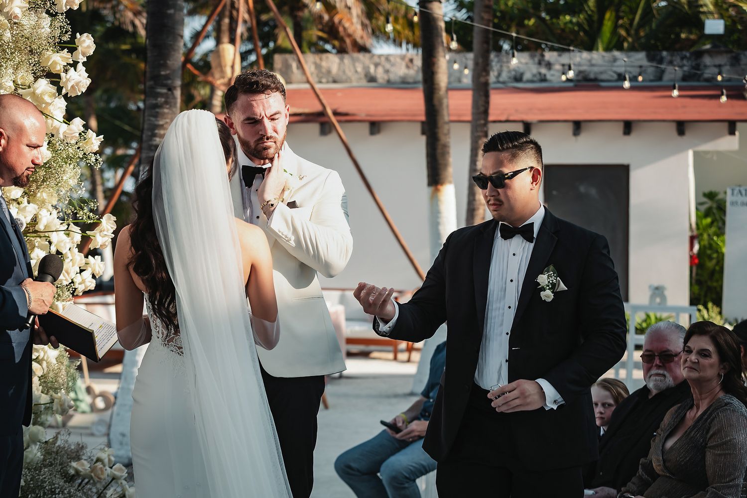 Outdoor wedding ceremony with groom in white tuxedo exchanging vows with bride in white gown and veil.
