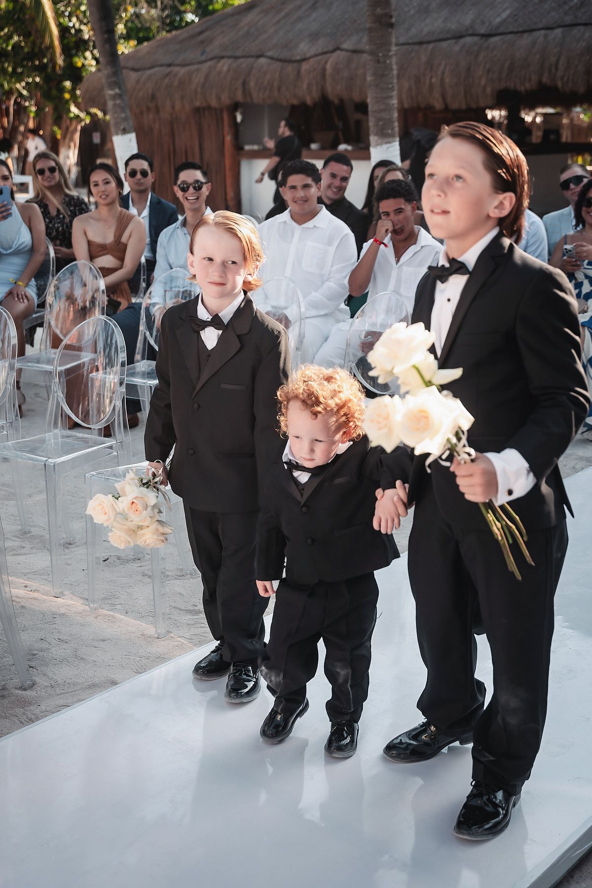 Three boys in black tuxedos walk down a white aisle holding white roses at an outdoor beach wedding ceremony.