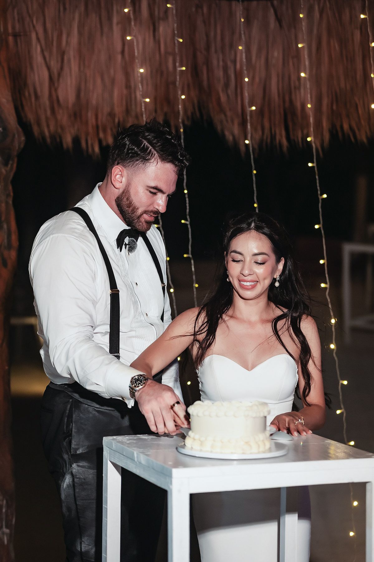 Bride and groom cutting their white wedding cake together under fairy lights at a tropical venue.