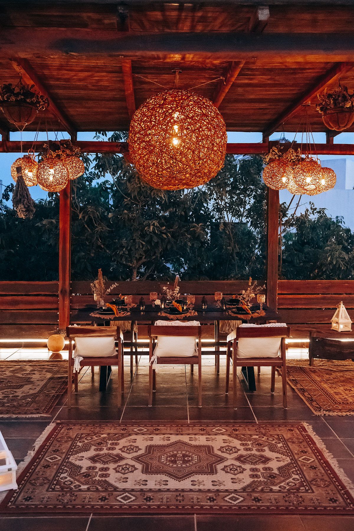 Cozy boho outdoor dining area at dusk with large woven globe pendant light, wooden chairs, and patterned rug.