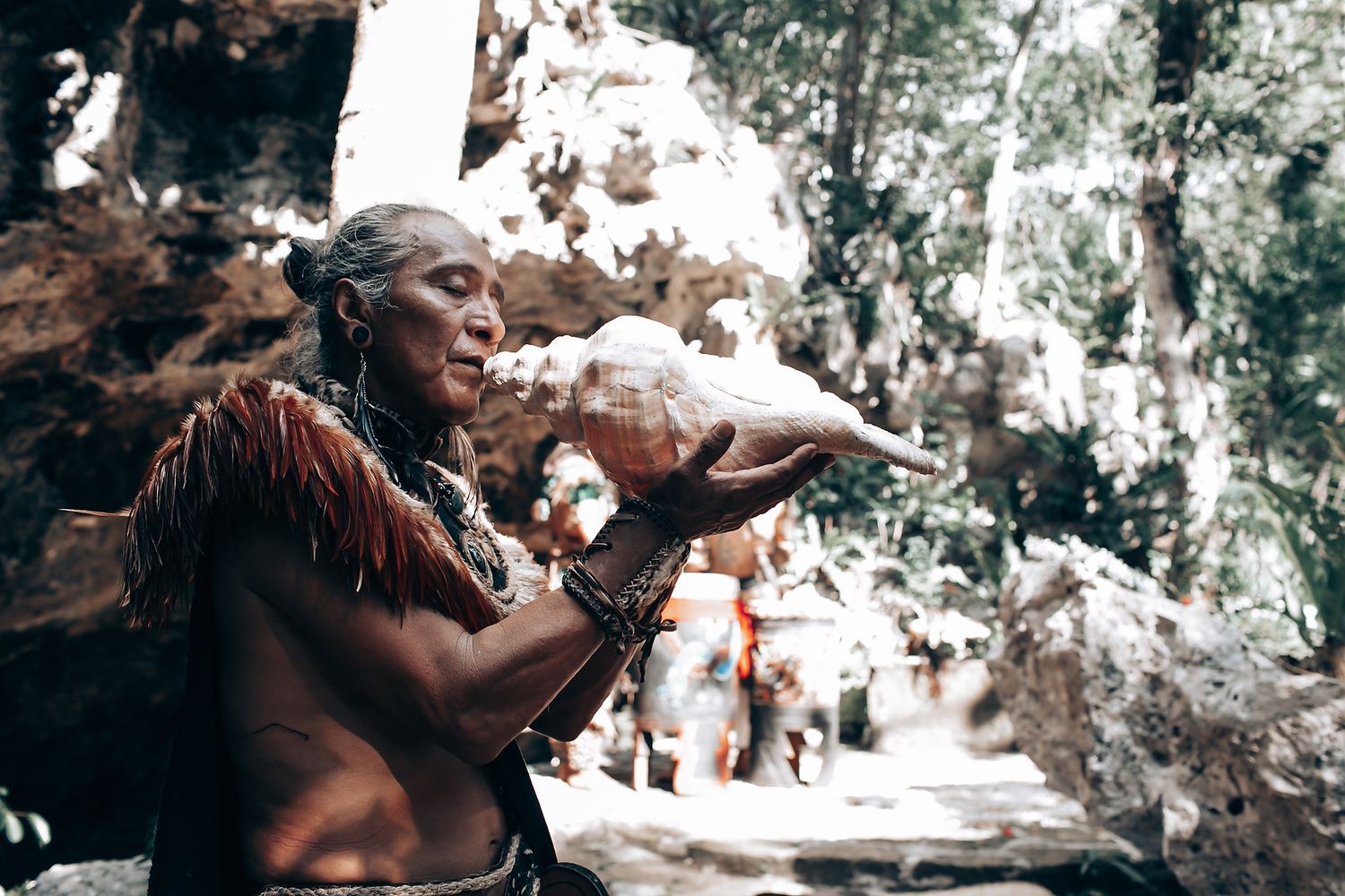 An elderly indigenous man blows a conch shell in a forest setting, wearing traditional feathered attire.