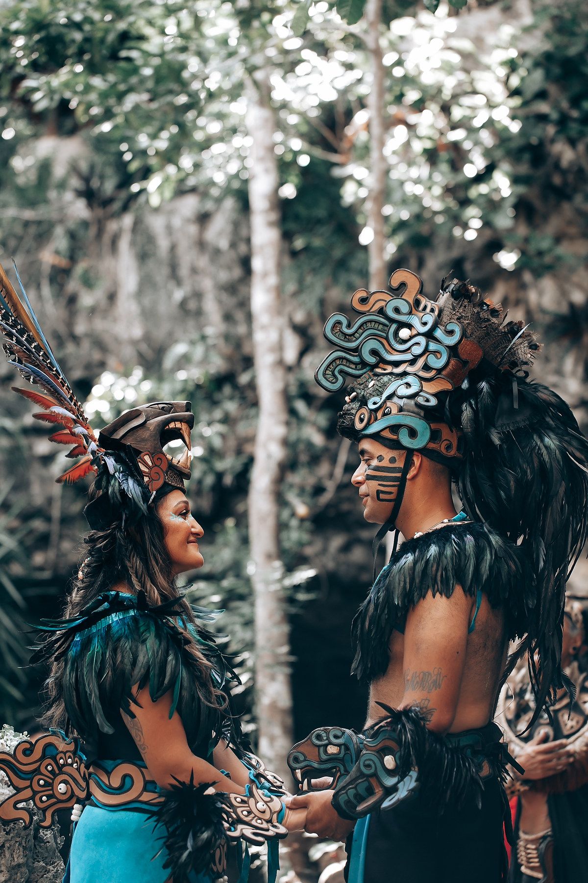 A couple in elaborate Mayan warrior costumes with feathered headdresses and face paint hold hands in a forest.