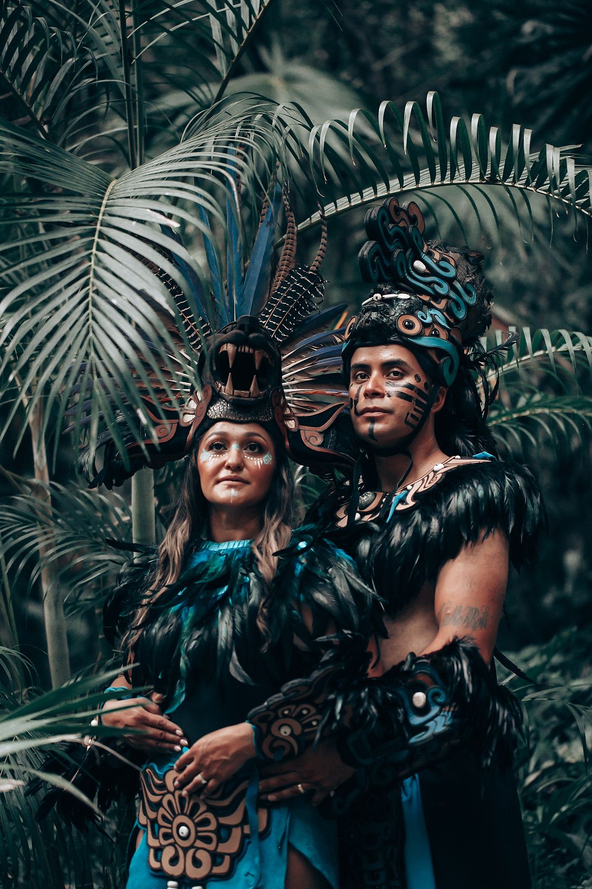Couple in elaborate Aztec feathered headdresses and dark ceremonial costumes pose before lush tropical palm fronds.