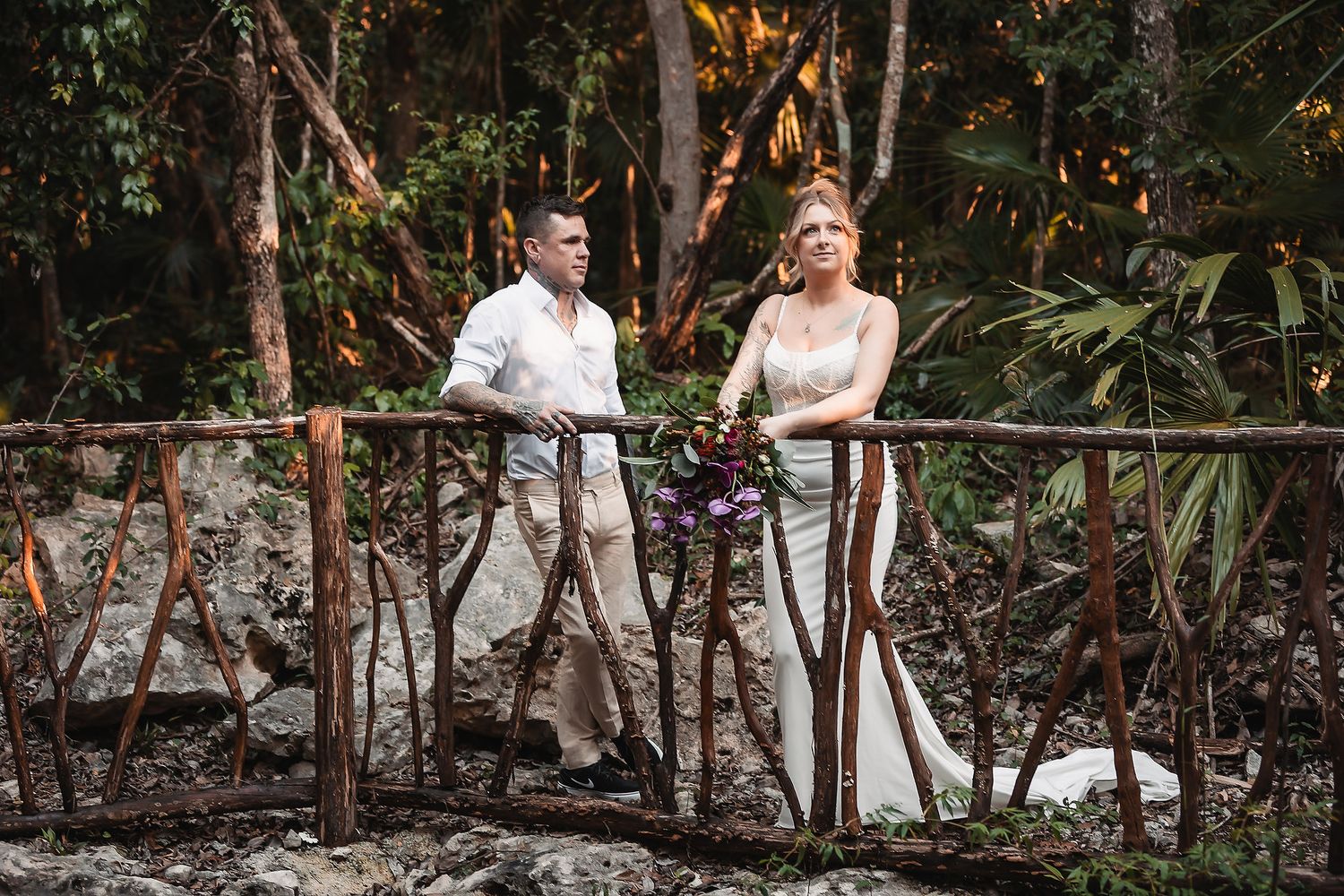 A couple poses on a rustic wooden railing surrounded by lush tropical foliage.