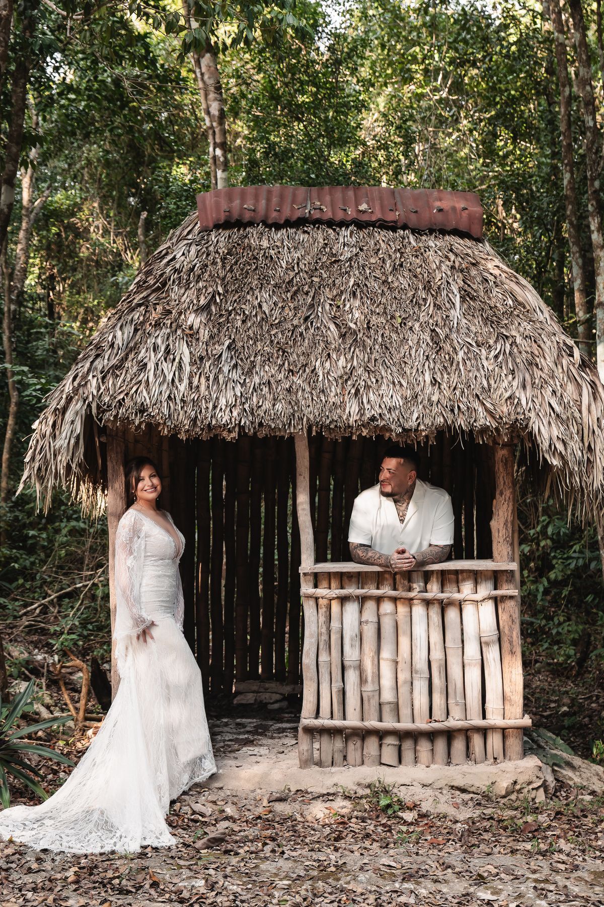 A couple poses at a rustic wooden kiosk with a thatched roof in a tropical forest setting.