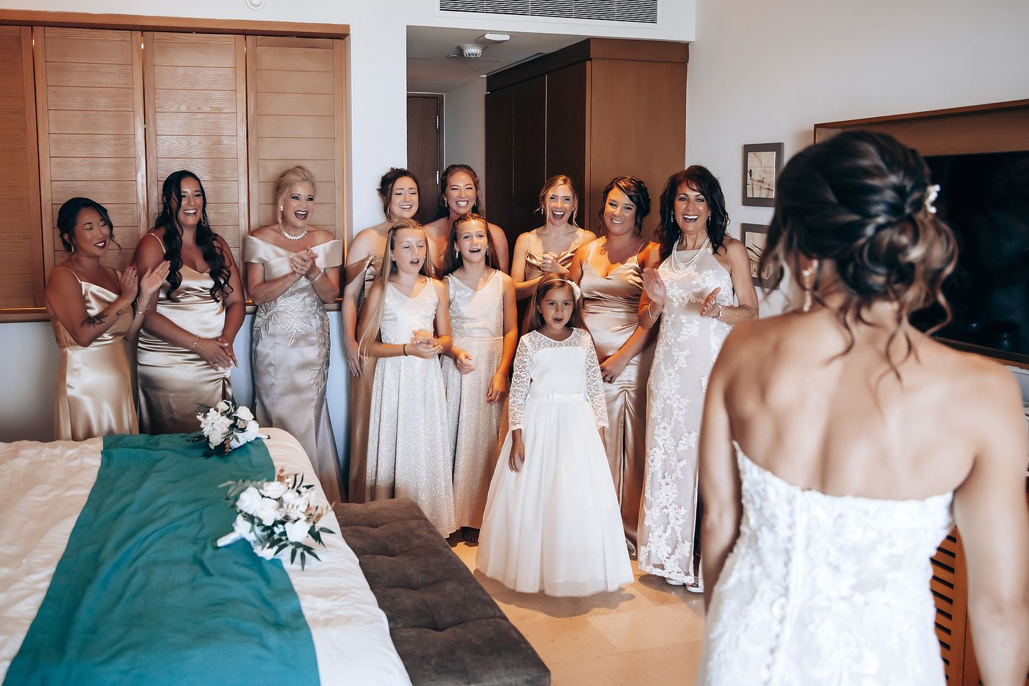 A group wearing white dresses pose together in a hotel hallway with matching bouquets.