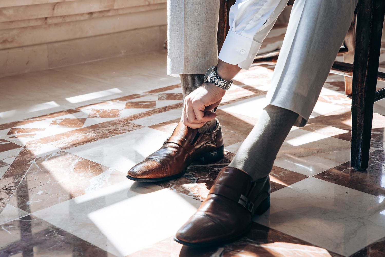 Brown leather dress shoes are shown on a person's feet while walking on a light wooden floor in sunlight.