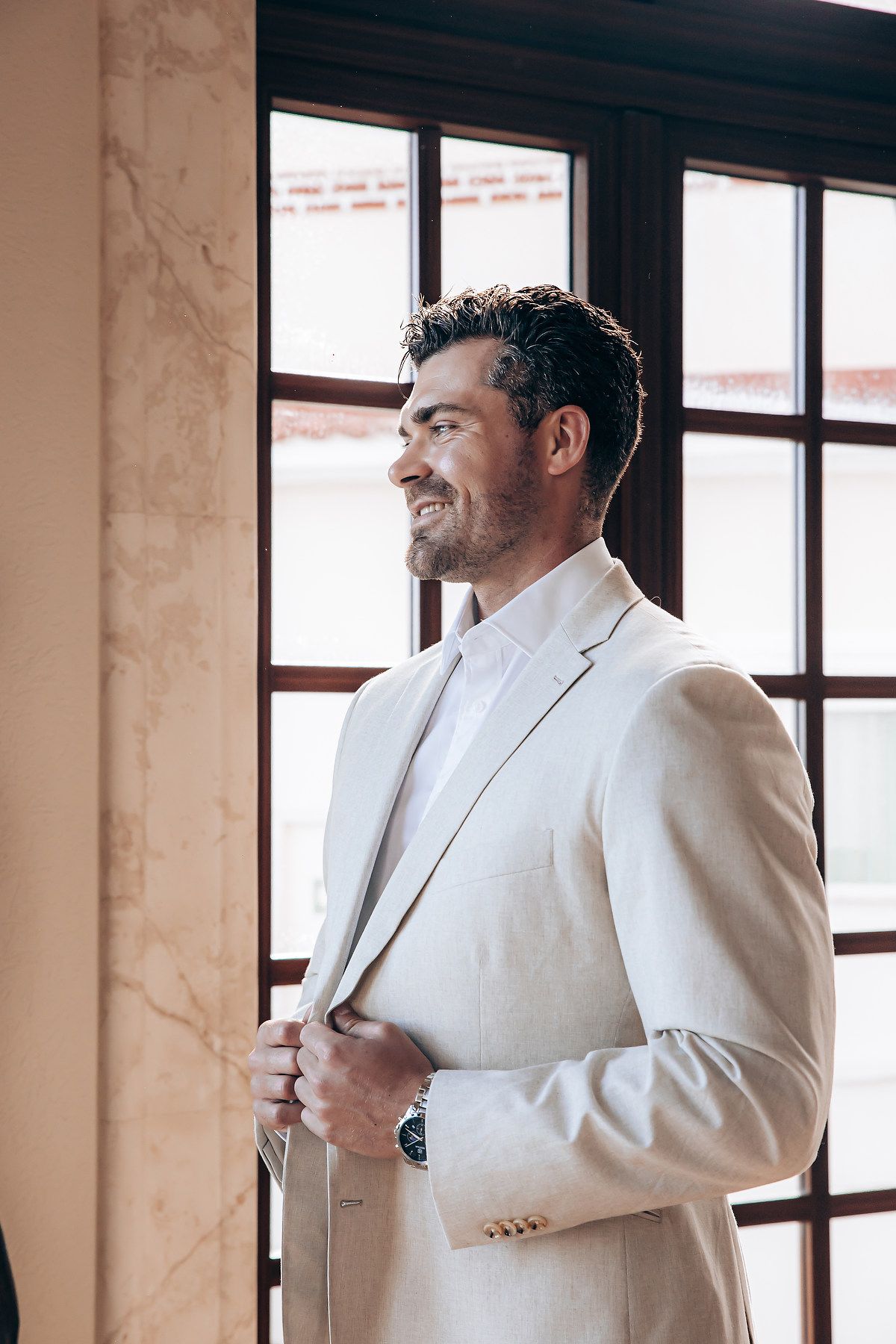 A person in a white suit jacket stands by a window, looking away from the camera in an elegant indoor setting.