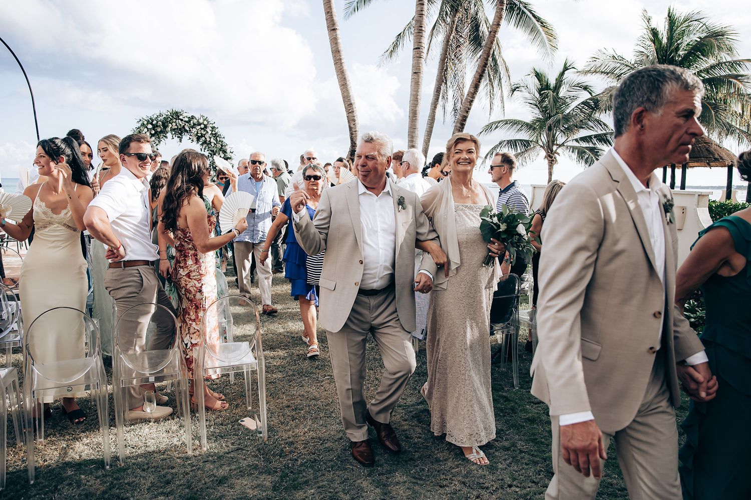 Wedding party in beige suits and dresses celebrate on a tropical beach with palm trees and ocean views.