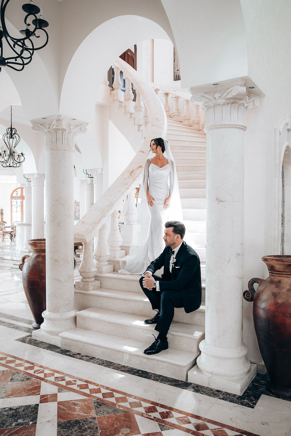 A couple in formal attire poses on an elegant white marble staircase with ornate columns and decorative urns.