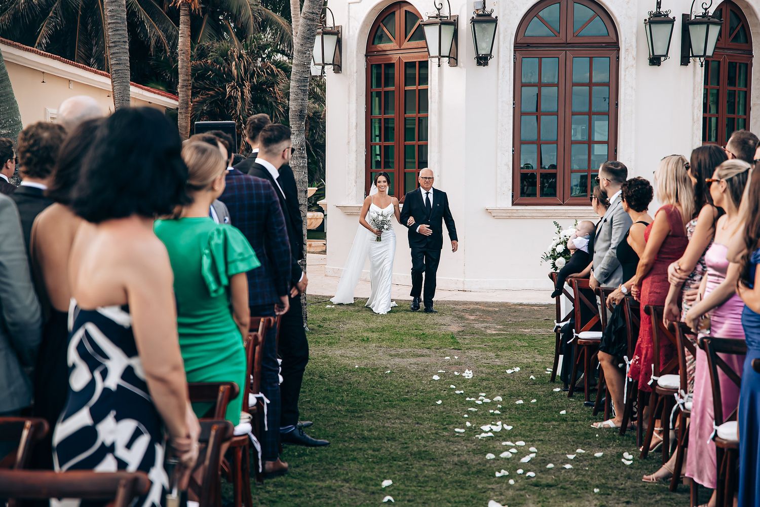 Wedding guests line up outdoors along a white stone church building with scattered flower petals on the grass.