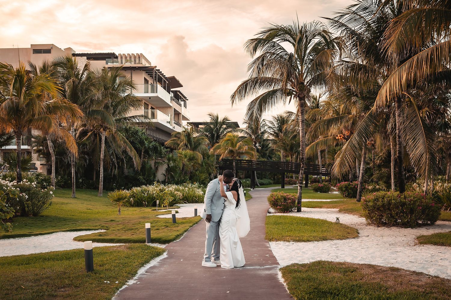 A couple walks along a palm-lined pathway at a tropical resort during sunset.