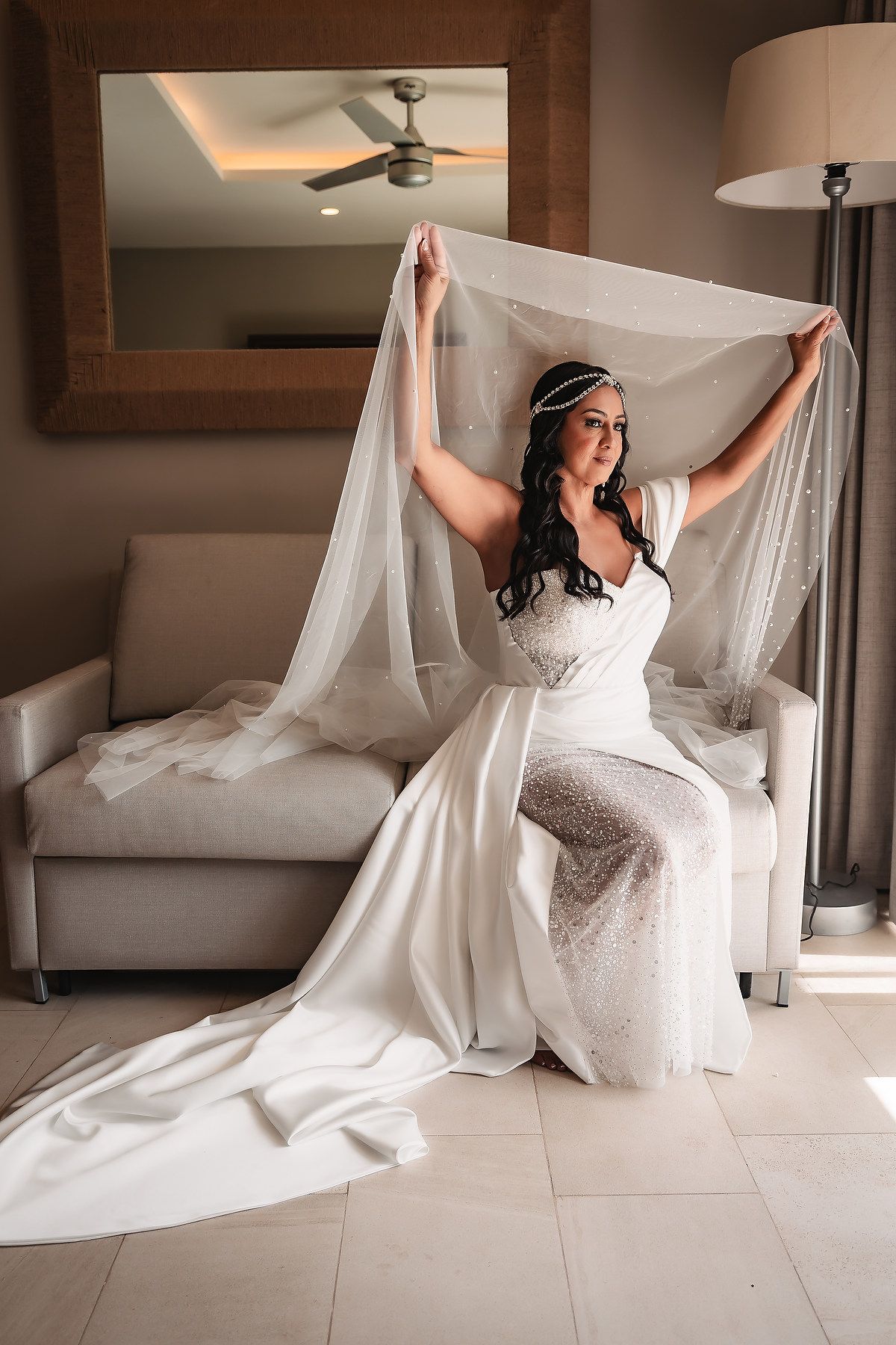 Bride in a white lace dress playfully poses with her veil in an elegant room.