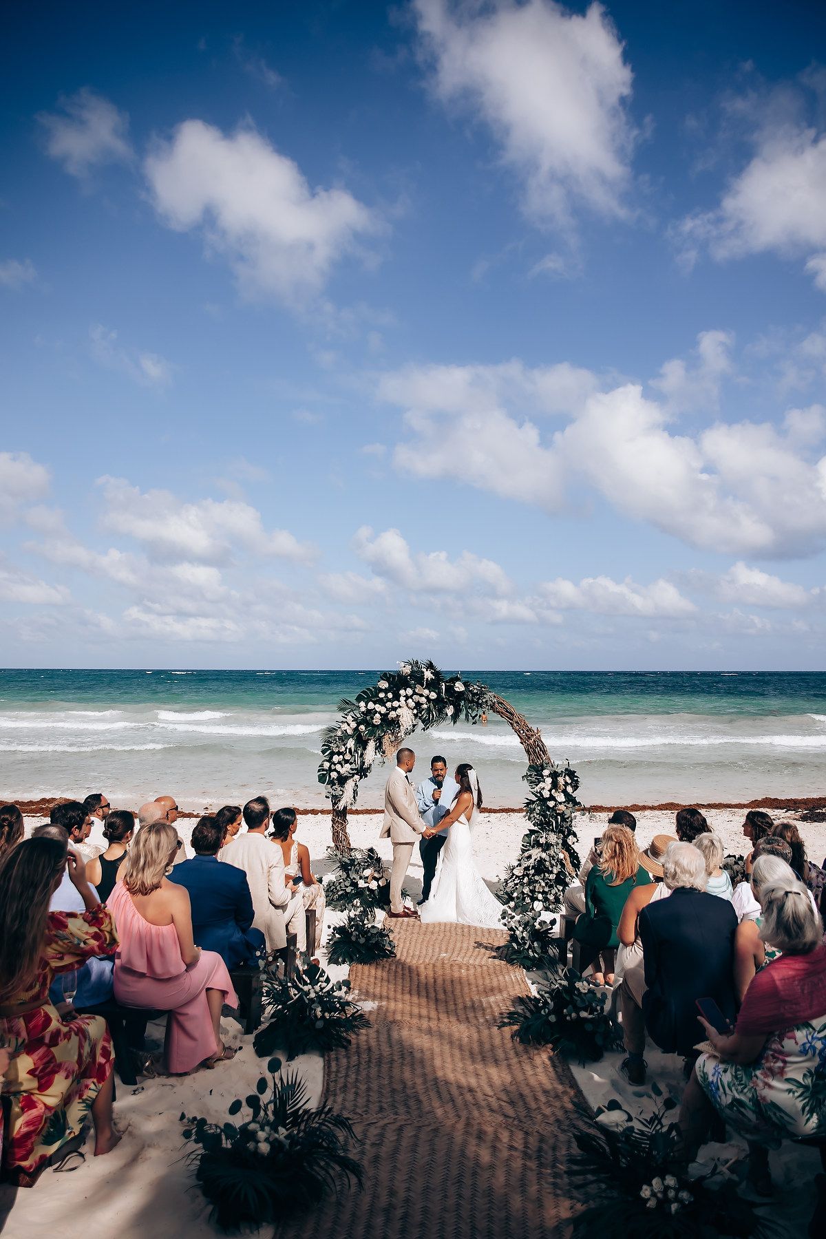 Scenic beach wedding setup with white floral arch against blue sky and rolling waves.
