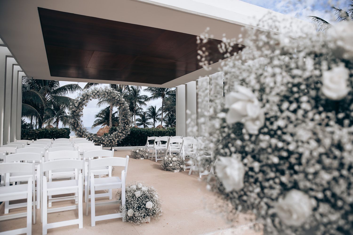 Outdoor wedding ceremony setup with white chairs and floral decorations under a bright sky.