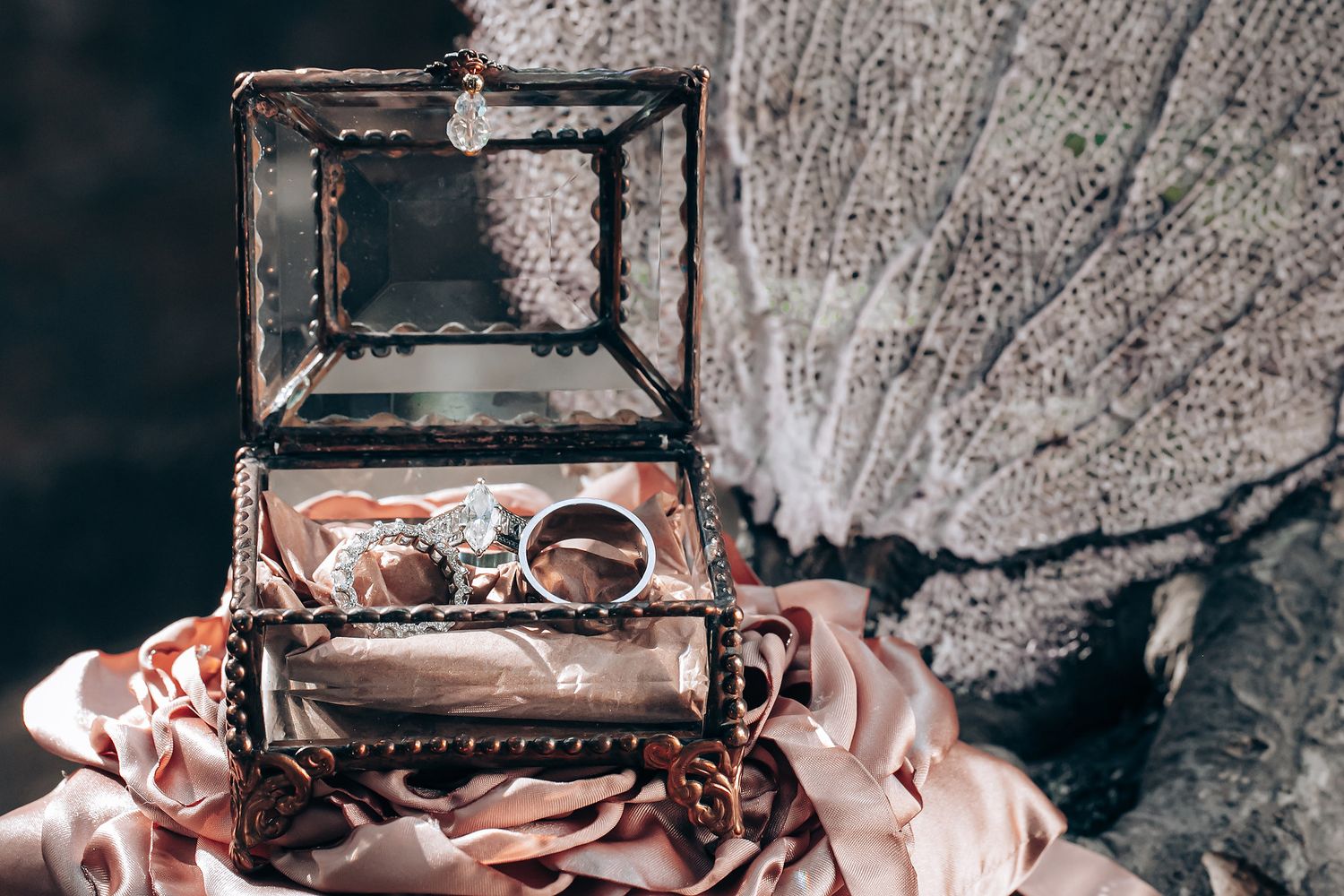 Wedding rings displayed in a vintage glass jewelry box with pink fabric and rustic textured background.