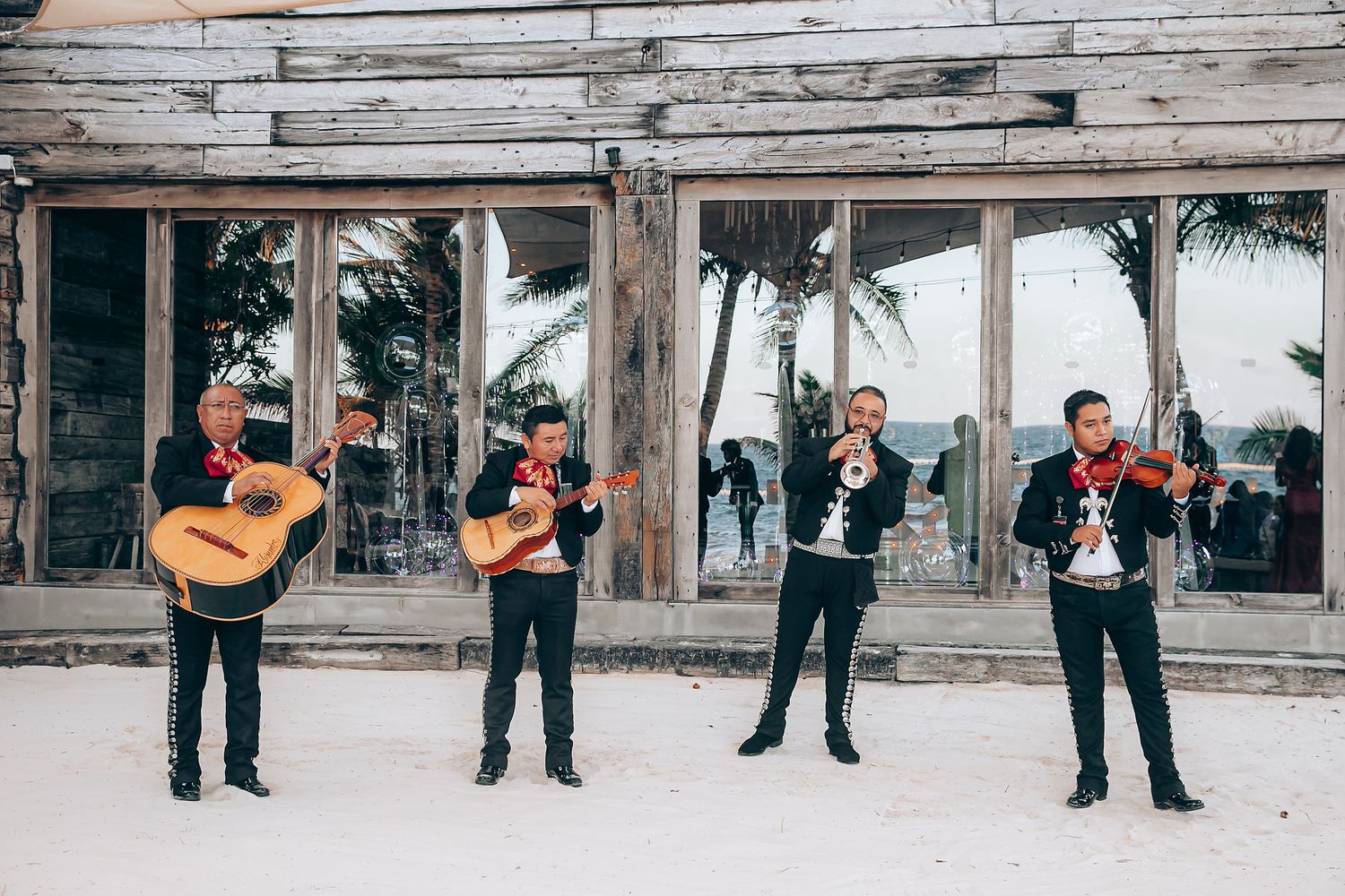 A mariachi band performs outdoors with guitars and trumpets on a beachfront wooden deck with palm trees in the background.