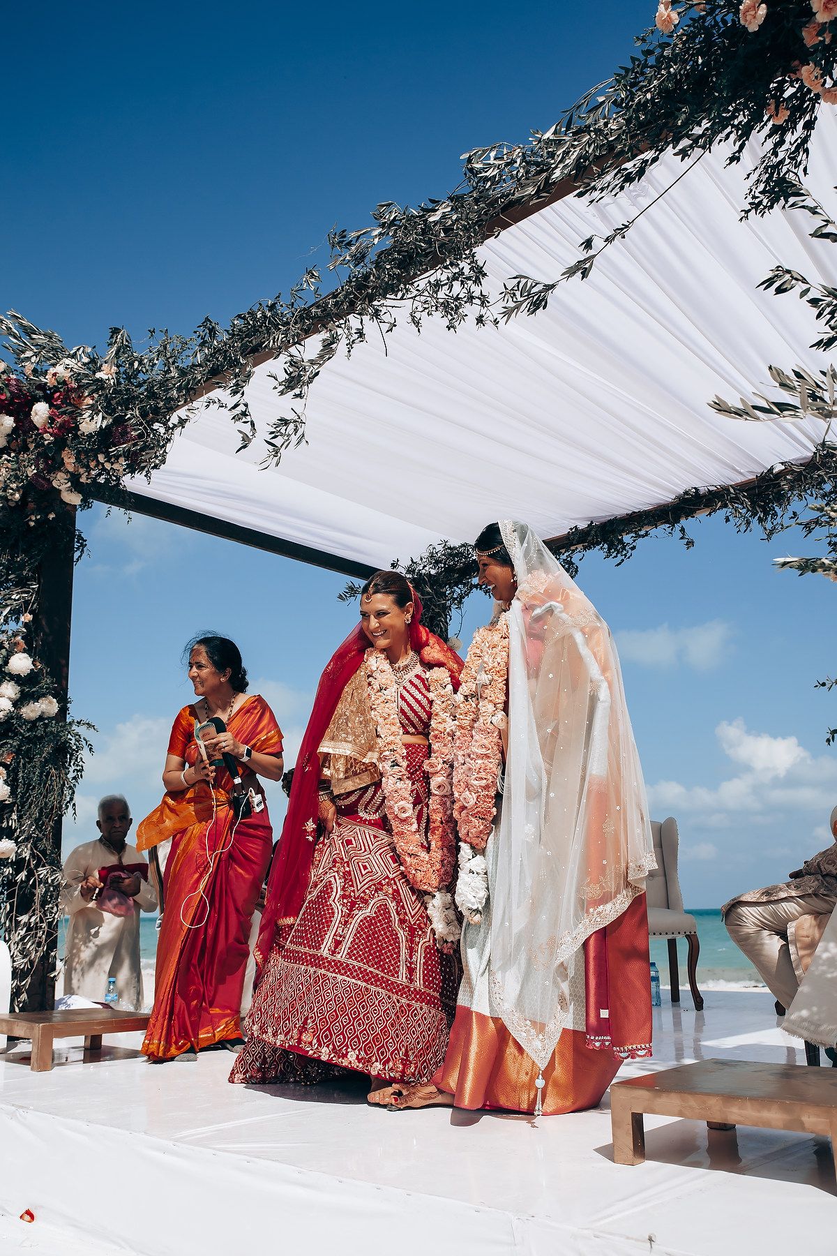 Beachfront Indian wedding ceremony with white draping and floral decorations against blue sky.