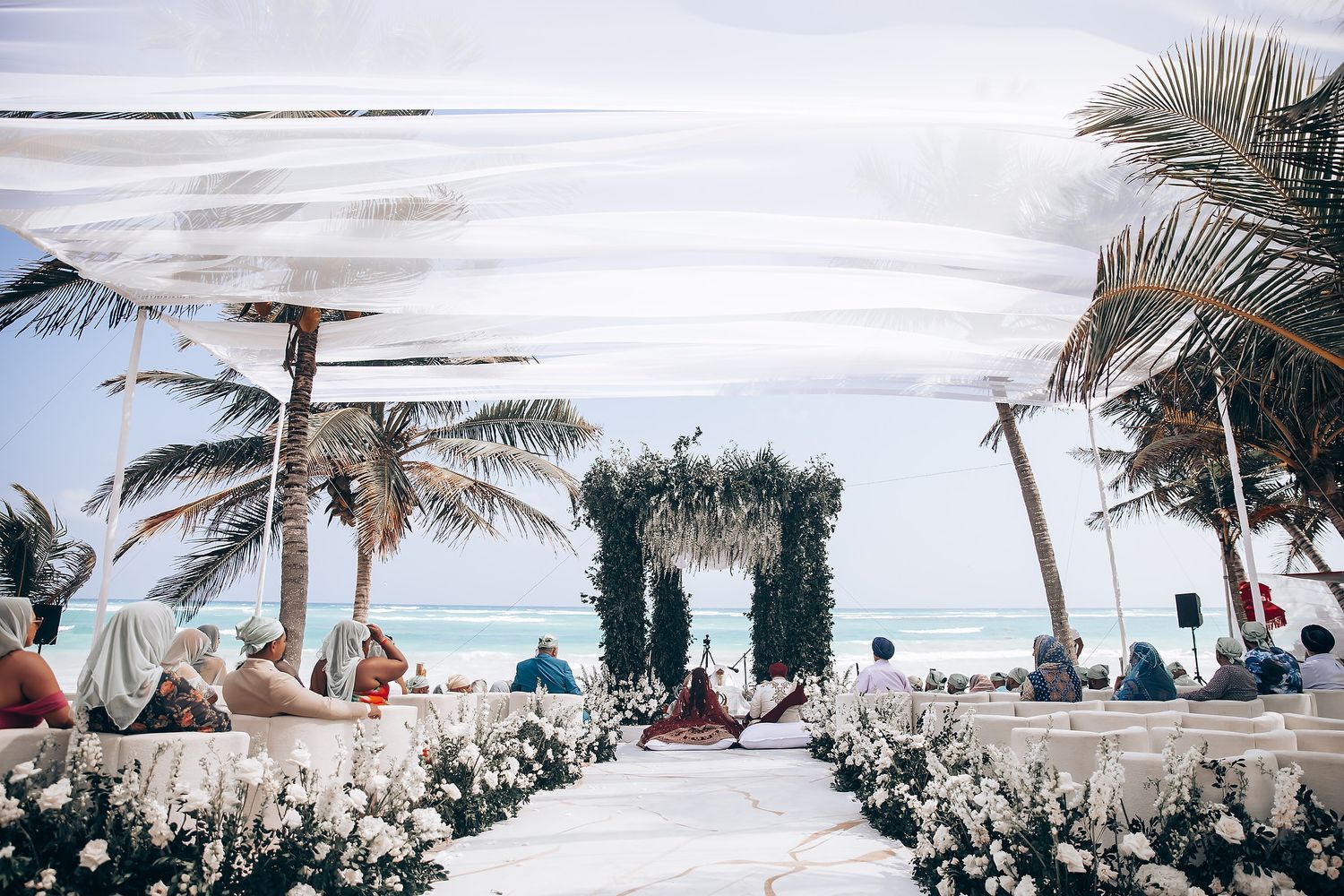 Beachfront wedding ceremony setup with white decor and natural palm tree backdrop.