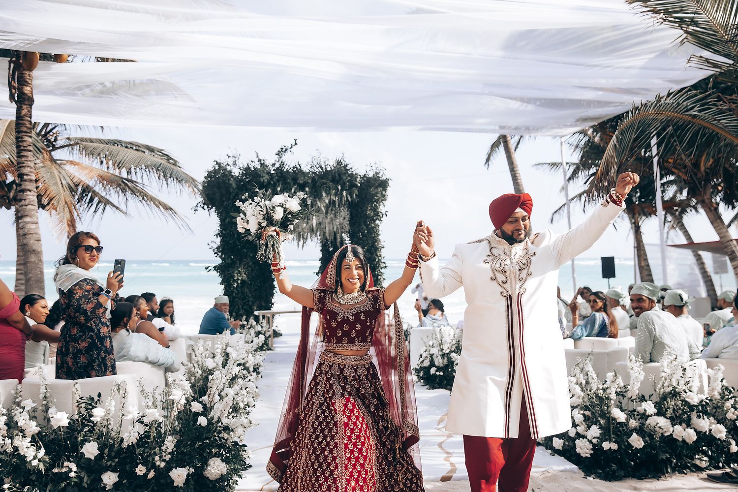 Tropical beach wedding setup with white chairs and floral arrangements.
