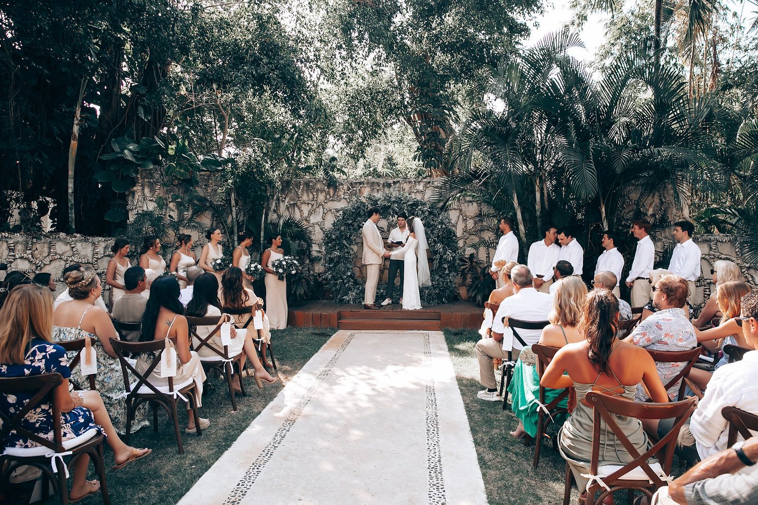 Wedding ceremony setup with guests seated outdoors under tropical trees.
